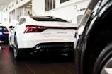 A sleek white Audi e-tron GT is prominently displayed in a well-lit car showroom. The car's modern design features bold, streamlined curves and distinctive rear lights. Other vehicles and modern architectural elements of the showroom are visible in the background.