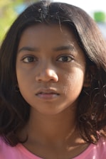A close-up portrait of a young girl with long, dark hair. Her expression is neutral, with a slightly focused gaze. The lighting creates soft shadows on her face, highlighting her features against a blurred natural background.