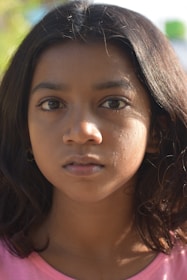 A close-up portrait of a young girl with long, dark hair. Her expression is neutral, with a slightly focused gaze. The lighting creates soft shadows on her face, highlighting her features against a blurred natural background.
