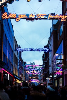 A busy urban street scene during evening time, featuring neon lights with lyrics from a famous song hanging overhead. The street is lined with shops and filled with a crowd of people.