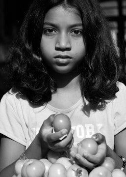 A young girl with medium-length hair is holding a few tomatoes in her hands. She has an expression of calm seriousness on her face and is wearing a T-shirt. The image is in black and white, providing a timeless and classic aesthetic.