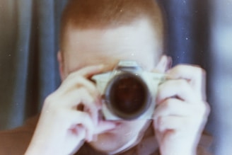 A person is holding a camera close to their face with both hands, seemingly capturing a photograph. The image is blurry with a vintage aesthetic and features soft, muted colors.