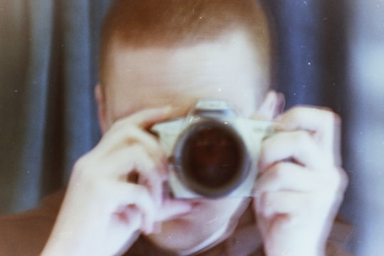 A person is holding a camera close to their face with both hands, seemingly capturing a photograph. The image is blurry with a vintage aesthetic and features soft, muted colors.