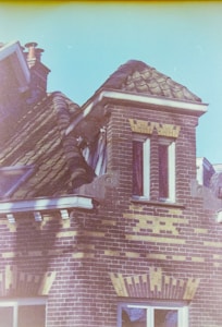 A detailed section of a traditional brick building with intricate brickwork patterns and a sloped tiled roof. The roof features a dormer window with white trim, and there's a visible chimney with a metal cap. The window panes are divided into smaller sections, emphasizing the architectural style.