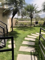 A small landscaped garden with neatly arranged stepping stones on a lush green lawn. Tall palm trees are scattered, providing shade and adding a tropical feel. The area is enclosed with black iron railings and a beige wall. In the background, distant hills are visible against a partly cloudy sky.