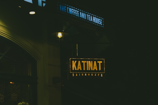 Dimly lit street scene featuring a brightly illuminated sign displaying the name 'KATINAT SaigonKafe', hanging under another sign that reads 'THE COFFEE AND TEA HOUSE'. The signs are the focal points, surrounded by darkness, implying nighttime.
