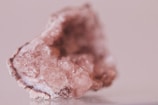 A close-up image of a rough, translucent pink mineral with crystalline structures. The stone's surface displays various facets and textures, with light reflecting off its shiny surfaces.