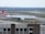 A large passenger airplane with the Turkish Airlines logo is positioned on an airport runway. The airplane is mostly white with red accents and is parked on a taxiway. The background features multiple runways and grassy fields, with some buildings visible in the distant background under a partly cloudy sky. The foreground shows a structure with windows and various airport equipment.