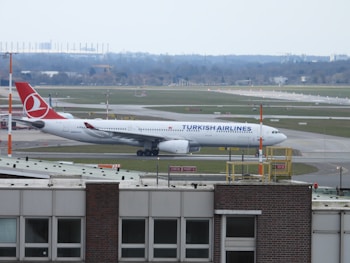 A large passenger airplane with the Turkish Airlines logo is positioned on an airport runway. The airplane is mostly white with red accents and is parked on a taxiway. The background features multiple runways and grassy fields, with some buildings visible in the distant background under a partly cloudy sky. The foreground shows a structure with windows and various airport equipment.