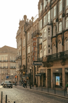 A charming European street lined with ornate historic buildings, featuring a prominent hotel sign. The architecture displays intricate details, large windows, and decorative balconies. Pedestrians and a car are visible, contributing to a lively urban atmosphere.