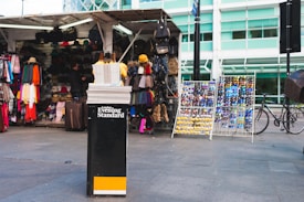 A street market stall displays various items including colorful scarves, hats, sunglasses, and bags. A stack of newspapers is prominently featured at the front labeled 'London Evening Standard'. Bicycles are visible on the right side, and the background consists of a modern building with glass windows.