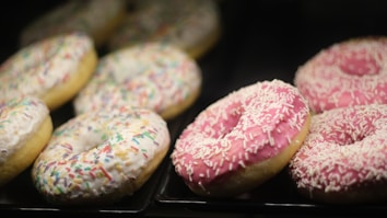 A selection of donuts with vibrant frosting and sprinkles appears on display. The donuts on the left are covered with white frosting and colorful sprinkles, while those on the right feature pink frosting with white sprinkles. They are arranged neatly on black trays, suggesting a bakery or cafe setting.