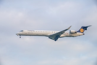 A commercial airplane with the Lufthansa logo is flying against a cloudy sky, displaying its white fuselage and distinctive tail fin.