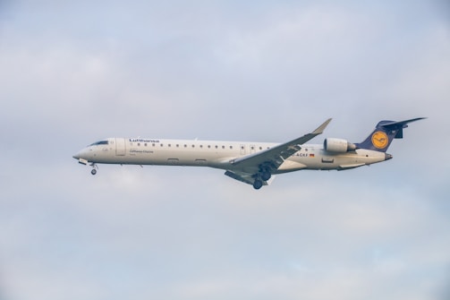 A commercial airplane with the Lufthansa logo is flying against a cloudy sky, displaying its white fuselage and distinctive tail fin.