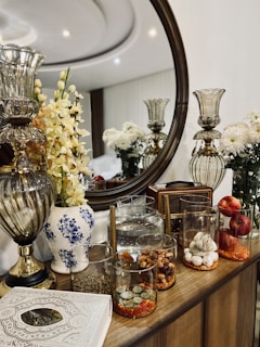 A beautifully arranged tabletop features a variety of decorative items, including a large round mirror, two ornate glass candle holders, and several vases with fresh white and yellow flowers. In front of the mirror, there is a retro-style radio, a water jug, and glass containers filled with assorted items like nuts, coins, and garlic bulbs. Additionally, two red apples add a touch of color.