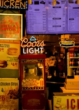 The scene features a vibrant food and beverage kiosk with colorful signage. Prominent neon signs for 'Coors Light' and 'Hot Dogs' stand out alongside menu boards advertising various food items such as popcorn, chicken wings, and soft drinks. A red popcorn machine is centrally located with a vintage design, and several advertisements for beverages, including Stella Artois and Coca-Cola, add to the bustling atmosphere. The lighting is warm and inviting, creating a classic fast-food or snack bar ambiance.