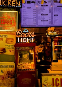 The scene features a vibrant food and beverage kiosk with colorful signage. Prominent neon signs for 'Coors Light' and 'Hot Dogs' stand out alongside menu boards advertising various food items such as popcorn, chicken wings, and soft drinks. A red popcorn machine is centrally located with a vintage design, and several advertisements for beverages, including Stella Artois and Coca-Cola, add to the bustling atmosphere. The lighting is warm and inviting, creating a classic fast-food or snack bar ambiance.