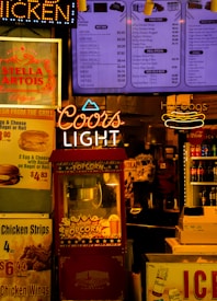 The scene features a vibrant food and beverage kiosk with colorful signage. Prominent neon signs for 'Coors Light' and 'Hot Dogs' stand out alongside menu boards advertising various food items such as popcorn, chicken wings, and soft drinks. A red popcorn machine is centrally located with a vintage design, and several advertisements for beverages, including Stella Artois and Coca-Cola, add to the bustling atmosphere. The lighting is warm and inviting, creating a classic fast-food or snack bar ambiance.