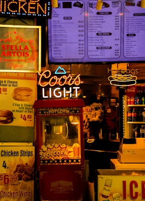 The scene features a vibrant food and beverage kiosk with colorful signage. Prominent neon signs for 'Coors Light' and 'Hot Dogs' stand out alongside menu boards advertising various food items such as popcorn, chicken wings, and soft drinks. A red popcorn machine is centrally located with a vintage design, and several advertisements for beverages, including Stella Artois and Coca-Cola, add to the bustling atmosphere. The lighting is warm and inviting, creating a classic fast-food or snack bar ambiance.