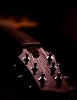A close-up view of a guitar headstock, featuring tuning pegs and strings, with a dark background highlighting the wood texture.