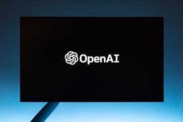 A black screen or display monitor with the OpenAI logo and text in white centered in the middle. The background is a gradient transitioning from dark to light blue from top to bottom.
