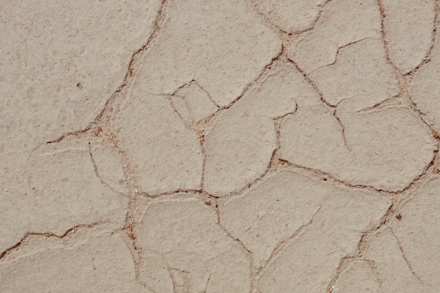 A close-up view of cracked, dry earth showing a pattern of irregular fissures and a predominantly dusty, beige surface.