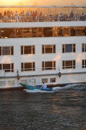 A large white cruise ship is moving through the water during sunset. There is a group of people standing on the upper deck, enjoying the view. Below the ship, a small boat with two people dressed in white is rowing alongside.
