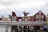 Several wooden houses painted in red and yellow are covered with snow. The buildings are situated along a waterfront, supported by stilts over water. A wooden walkway or bridge runs in front, and seagulls are flying in the cloudy, overcast sky.