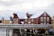 Several wooden houses painted in red and yellow are covered with snow. The buildings are situated along a waterfront, supported by stilts over water. A wooden walkway or bridge runs in front, and seagulls are flying in the cloudy, overcast sky.