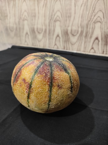 A round, textured fruit with a mottled surface of yellow, orange, red, and green streaks is placed on a black fabric with a wood-patterned background.