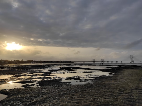 The image captures a serene coastal landscape during sunset, with a prominent bridge extending across the horizon. The sky is partly cloudy, with the sun peeking through, casting a warm glow over the scene. Tidal pools are visible on the shore, reflecting the sky above.