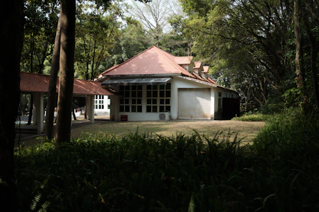 A house with a red-tiled roof is surrounded by trees and foliage. The building features multiple windows and a closed garage door. The area is shaded by tall trees, and the ground is covered with grass and plants.
