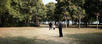 A park setting with a path surrounded by tall trees. Two adults and a child are walking along the path, enjoying the outdoors. Another individual stands off to the side, observing the scene. The trees create a natural canopy, and light filters through the leaves.