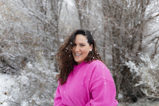 A woman wearing a bright pink sweater stands outside in a snowy landscape, with snowflakes gently falling around her. She has long, curly hair and is smiling warmly, creating a contrast between her vibrant attire and the muted, wintry background of leafless trees and snow-covered ground.