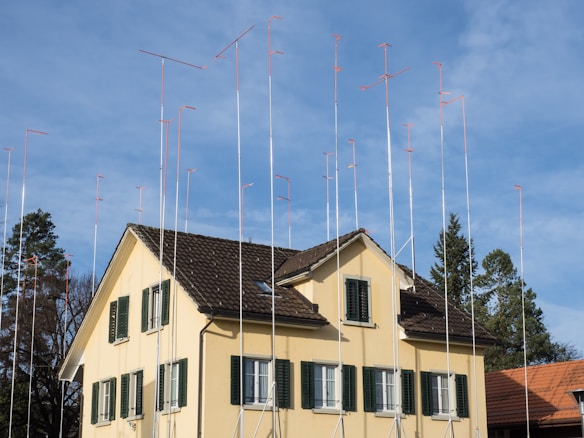 A house with a tiled, dark brown roof and yellow walls is surrounded by numerous tall metal poles, each with a small red-orange horizontal piece on top. The house has several green window shutters and is set against a clear blue sky. In the background, there are a few trees visible.
