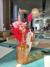 A table is set with a textured teal placemat on top of which sits a small amber glass vase containing dried pink and red flowers. In the background, a modern dining area is visible with white furniture and ambient lighting.