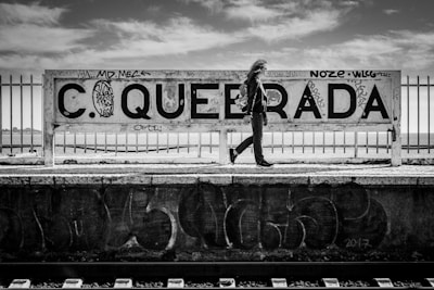 A person with long hair is walking alongside a graffiti-covered sign that reads 'QUEBRADA' at a train station. The station platform has graffiti, and metal fencing is visible behind the sign. The sky is partly cloudy, and the scene is in black and white.