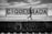 A person with long hair is walking alongside a graffiti-covered sign that reads 'QUEBRADA' at a train station. The station platform has graffiti, and metal fencing is visible behind the sign. The sky is partly cloudy, and the scene is in black and white.