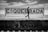 A person with long hair is walking alongside a graffiti-covered sign that reads 'QUEBRADA' at a train station. The station platform has graffiti, and metal fencing is visible behind the sign. The sky is partly cloudy, and the scene is in black and white.