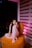 A young girl with long hair sits cross-legged on an orange chair, dressed in a light pink outfit. She rests her head on her hand, looking directly at the camera. The room is dimly lit with a modern curtain behind her and a soft, purple light casting a glow.