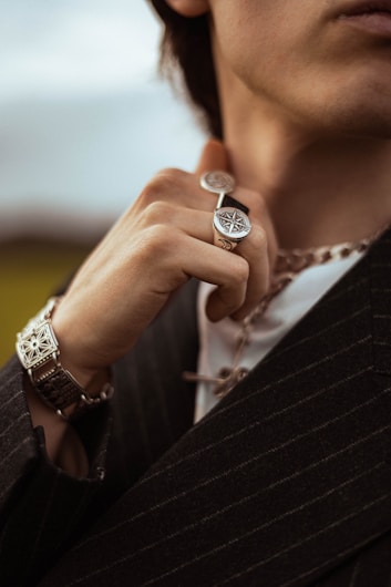 A close-up view of a person wearing jewelry, including a detailed ring with a compass design and a thick chain necklace. The person is dressed in a striped jacket, holding the ring near their face, with a bracelet visible on their wrist.