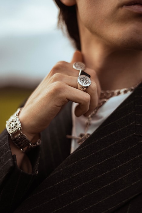 A close-up view of a person wearing jewelry, including a detailed ring with a compass design and a thick chain necklace. The person is dressed in a striped jacket, holding the ring near their face, with a bracelet visible on their wrist.