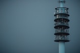 A tall, modern communication tower stands against a muted gray-blue sky. The structure is cylindrical with multiple levels, each adorned with various antennas and satellite dishes. Its sleek design and lights indicate it is active and operational.