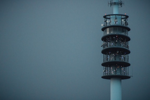 A tall, modern communication tower stands against a muted gray-blue sky. The structure is cylindrical with multiple levels, each adorned with various antennas and satellite dishes. Its sleek design and lights indicate it is active and operational.
