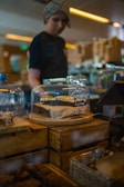 A bakery display features a ginger and lime cake priced at £3.50, placed under a clear glass dome on a wooden crate. In the background is a person wearing a headscarf, standing near other food items and bakery displays in a cozy, warm-lit environment.