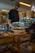 A bakery display features a ginger and lime cake priced at £3.50, placed under a clear glass dome on a wooden crate. In the background is a person wearing a headscarf, standing near other food items and bakery displays in a cozy, warm-lit environment.