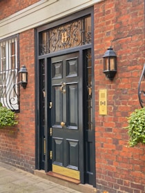 A dark, ornate wooden front door with brass accents is set in a traditional red brick building. Decorative ironwork is present above the door, featuring a number. There are two wall-mounted lanterns on either side of the door and window bars with intricate designs on a nearby window. Potted plants with green foliage add a touch of nature to the scene.