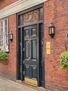 A dark, ornate wooden front door with brass accents is set in a traditional red brick building. Decorative ironwork is present above the door, featuring a number. There are two wall-mounted lanterns on either side of the door and window bars with intricate designs on a nearby window. Potted plants with green foliage add a touch of nature to the scene.