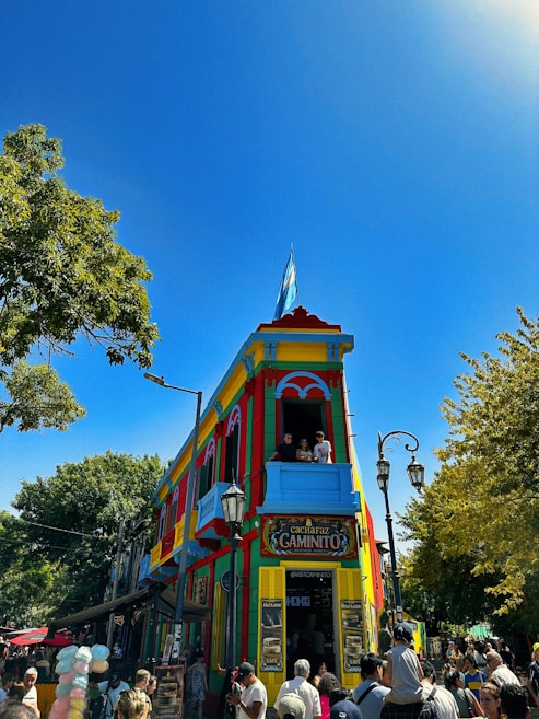 A vibrant, colorful building with bright blue, red, green, and yellow hues stands prominently. It has an Argentine flag on the roof and a sign reading 'Caminito'. People are gathered around the building, some on the balcony and others below. The sky is clear and blue, with trees adding greenery to the scene.