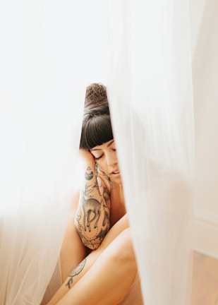 A person with dark hair and tattoos adorns their arm, sitting with a serene expression behind sheer white curtains. The soft lighting creates a gentle, dreamy atmosphere.